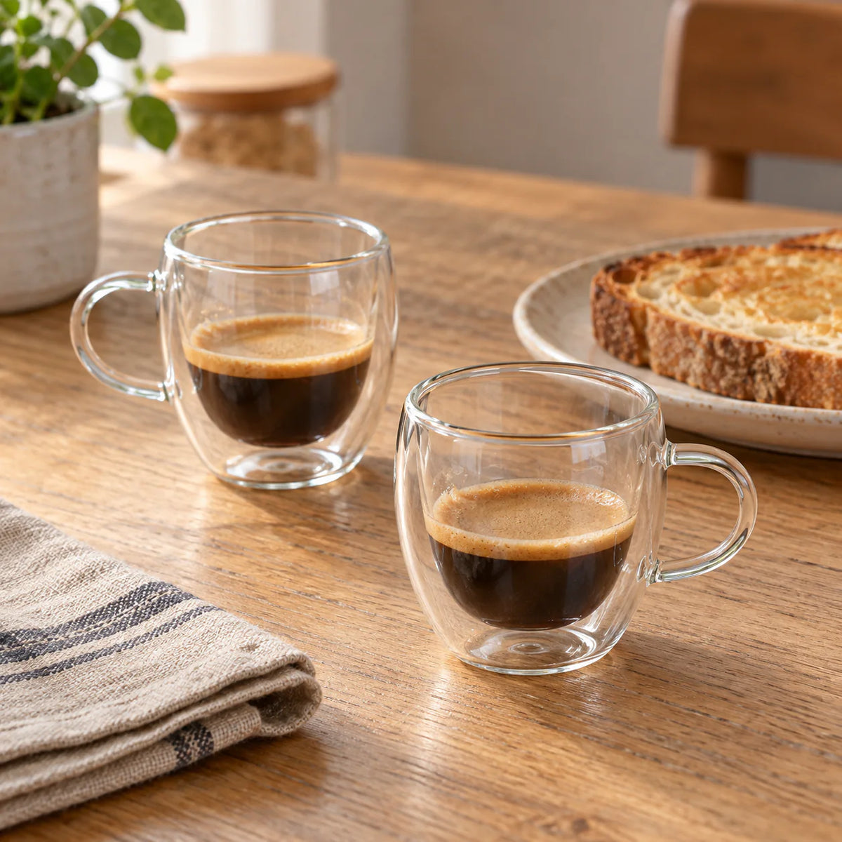 Duas xícaras de vidro com alça e espresso em mesa de café da manhã com pão e guardanapo