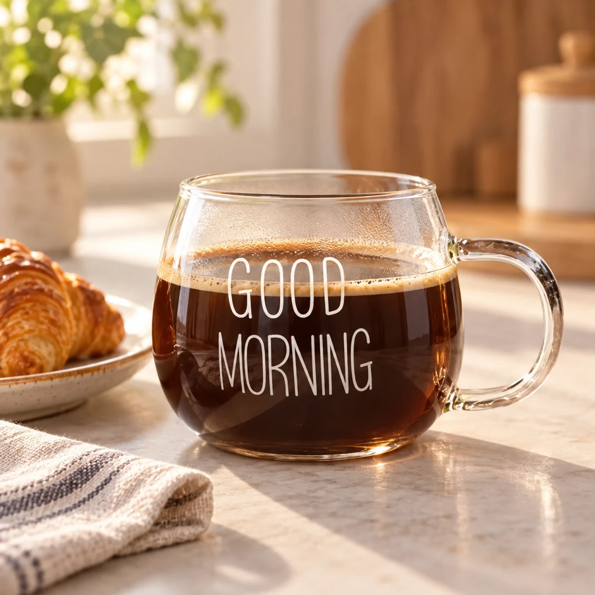 Xícara de vidro Good Morning Lumi Homi com estampa branca e café, ao lado de um croissant sobre a mesa